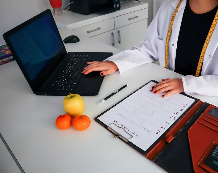 A person in a white coat sits at a desk with a laptop, a clipboard with a weekly plan, an apple, and two tangerines, conveying a professional, organized tone.