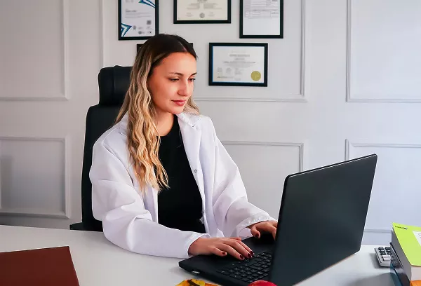 A woman in a white lab coat sits at a desk working on a laptop in a bright office, with framed certificates on the wall behind her