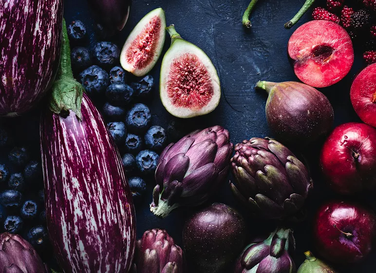 A vibrant assortment of purple vegetables and fruits, including striped eggplants, figs, blueberries, artichokes, plums, and apples, on a dark surface.