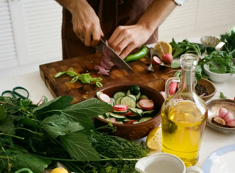 A person chops vegetables on a wooden board, surrounded by fresh greens, radishes, cucumbers, and a bottle of olive oil, conveying a fresh, healthy vibe.