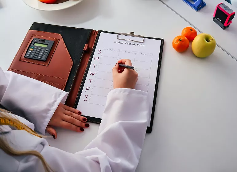 A person in a lab coat writes on a weekly meal plan clipboard, surrounded by a calculator, apples, and oranges. The setting is organized and professional.