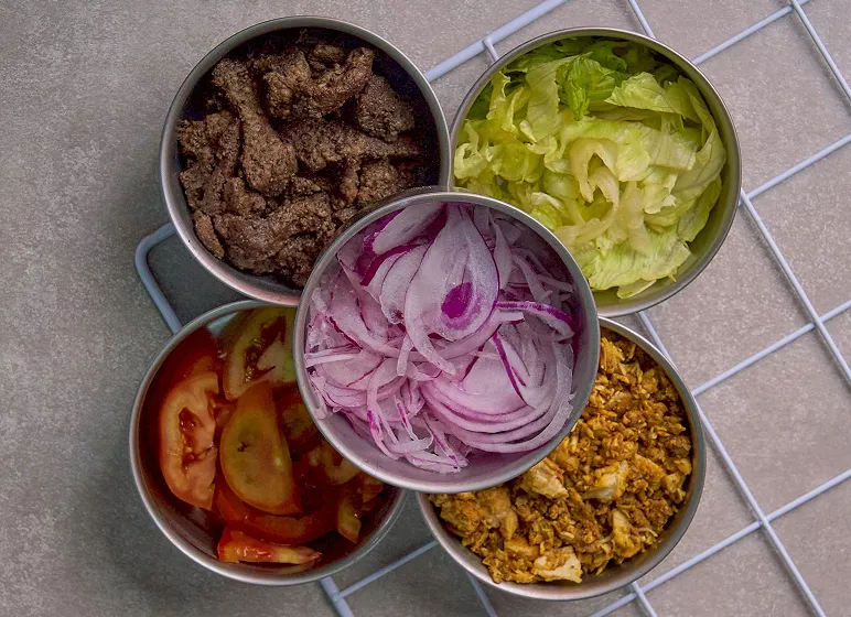 Five metal bowls with sliced onions, lettuce, tomato slices, cooked beef, and crumbled egg on a gray surface. The arrangement is neat and colorful.