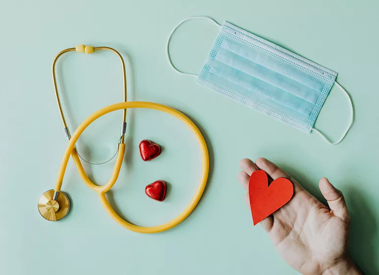 Yellow stethoscope and two heart-shaped candies on a light green background. A hand holds a red paper heart, with a blue surgical mask nearby, conveying care.