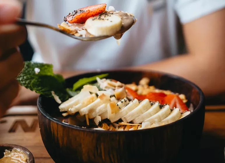 A wooden bowl filled with a vibrant smoothie topped with sliced bananas, strawberries, coconut, chia seeds, and mint. A spoonful of toppings is held above, evoking freshness and health.