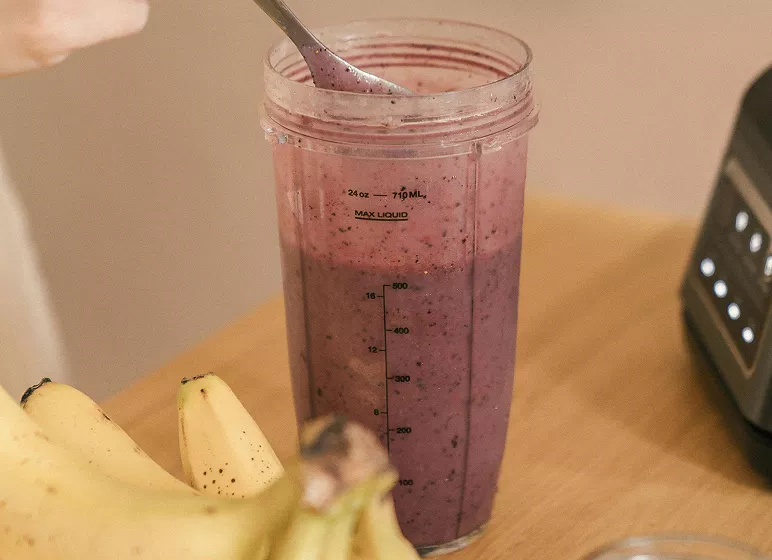 A tall blender cup filled with a purple smoothie, stirred by a spoon. Bananas are in the foreground. The scene suggests a healthy, fresh breakfast.
