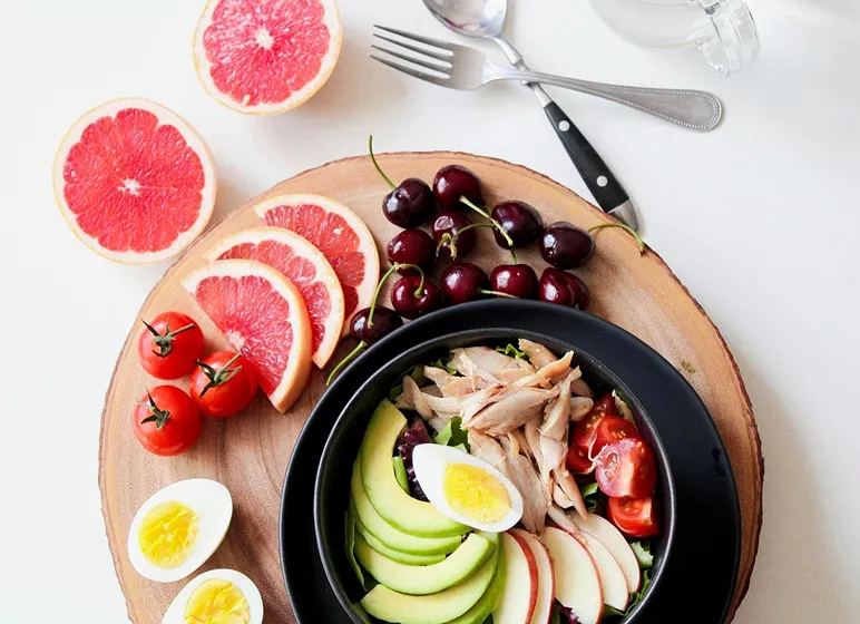 A vibrant, healthy meal is arranged on a wooden platter featuring sliced grapefruit, cherries, cherry tomatoes, and a salad bowl with avocado, eggs, and chicken.
