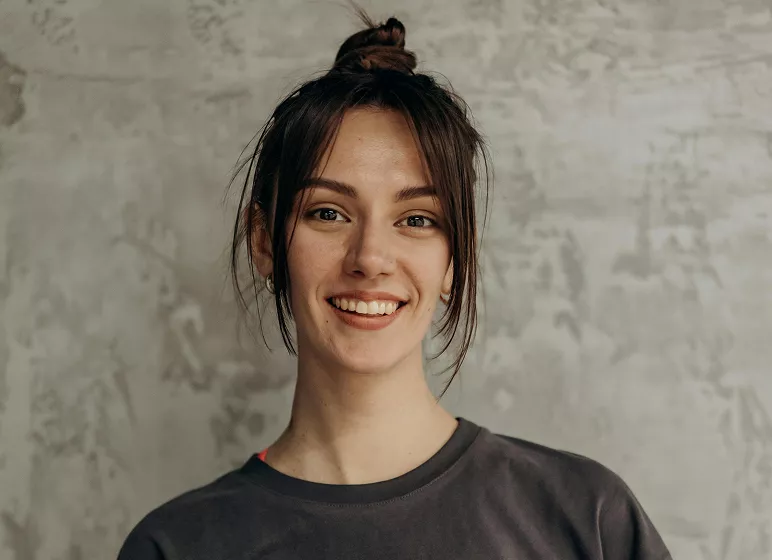 Smiling person with dark hair in a messy bun wearing a dark shirt against a textured gray background, conveying a cheerful and relaxed vibe.