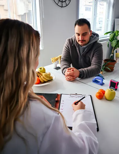 Nutritionist reviewing a meal plan with a man seated at a table, with fruits, a clipboard, and healthy food items visible in a bright kitchen setting.