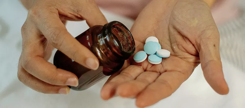 Hands pouring blue and white pills from a brown bottle into one palm, possibly depicting medication handling or healthcare. Background is softly blurred.
